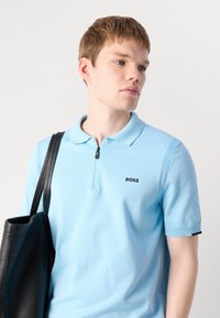 Polo azul claro com gola de fecho, mangas curtas e detalhes texturizados. Apresenta um logótipo preto no peito e um design discreto e moderno.