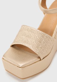 Sandale plateforme dorée avec une tige en cuir synthétique texturé, dotée d'une large bride avec un motif en relief et une finition légèrement brillante.