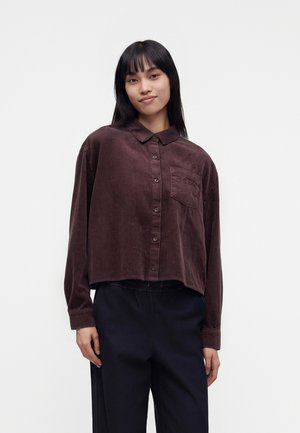 Joven con el pelo liso y negro, lleva una camisa de pana marrón oscuro y suelta con botones y pantalones negros, de pie frente a un fondo sencillo.
