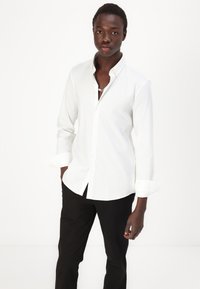 Cămașă albă cu mânecă lungă, cu guler butonat și mâneci răsucite, combinată cu pantaloni negri. Material din bumbac neted, fără modele vizibile.
