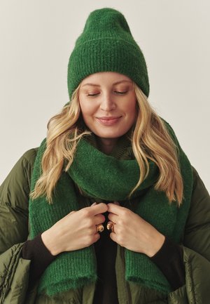 Grøn strikket hue og matchende tørklæde lavet af blødt materiale. Tørklædet har en tekstureret overflade og bæres med en grøn dunjakke over en sort sweater.
