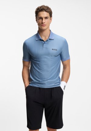 Mann trägt hellblaues BOSS-Poloshirt und schwarze Shorts, mit weißem Handgelenkband am linken Handgelenk, steht mit den Händen in den Taschen.