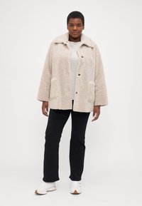 Beige, fluffy shearling jacket with a notched collar, front pockets, and black buttons. Worn over a light gray top and black pants.