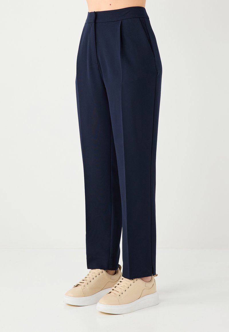 Pantalon marine taille haute, avec un devant plissé et des jambes droites. Confectionné en tissu lisse, porté avec des baskets beige clair.