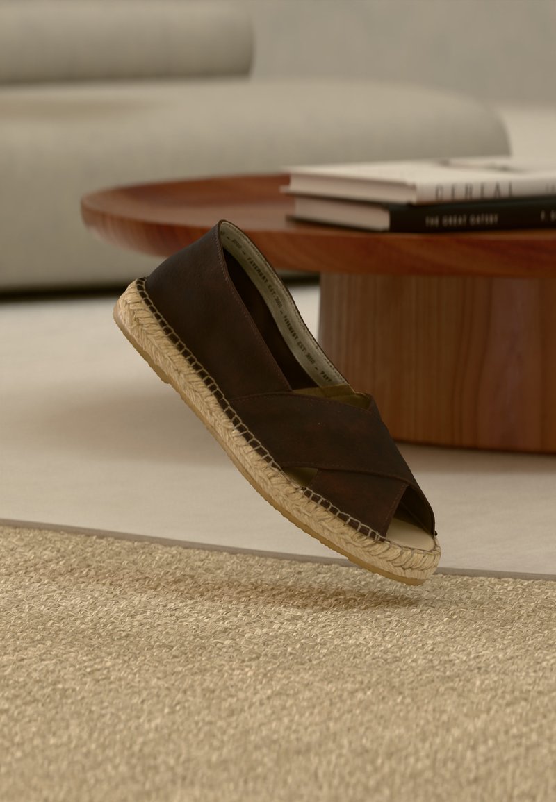 Chaussure espadrille marron foncé avec semelle tissée flottant au-dessus d'un tapis beige texturé, table basse en bois et livres empilés en arrière-plan.