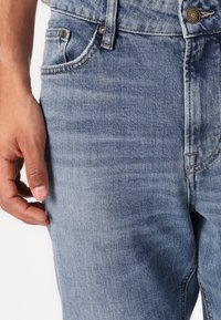Blugi din denim albastru deschis cu o textură subtilă, design cu cinci buzunare și închidere cu un nasture metalic la talie. Mână odihnindu-se pe lateral.
