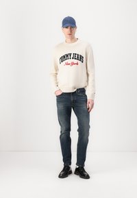 Pulover crem cu inscripția "TOMMY JEANS New York" în litere bleumarin și roșii, asortat cu blugi închis la culoare și pantofi negri, purtat cu o pălărie albastră.