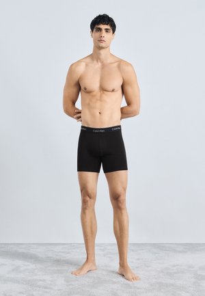 Boxer in cotone neri con un elastico con logo, realizzati in un materiale liscio e morbido. Il modello è in piedi a piedi nudi su una superficie grigio chiaro.