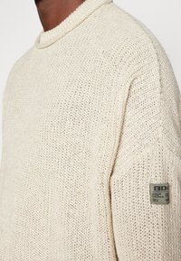 Cremfarbener gestrickter Pullover mit strukturierter Oberfläche, der einen runden Ausschnitt und ein kleines Stoffetikett am Ärmel aufweist.