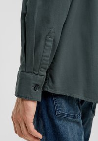 Nahaufnahme eines dunkelgrünen Langarmhemdmanschette mit Knopfdetail, getragen über blauen Jeans.