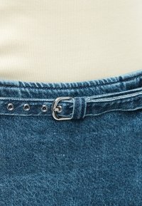 Κοντινή λήψη μπλε ζώνης από τζιν με ασημένια αγκράφα, δεμένη γύρω από τη μέση μπλε τζιν παντελονιού, συνδυασμένη με μπλούζα ριπ σε εκρού απόχρωση.