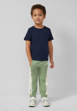 Kind steht mit Händen in den Taschen, trägt ein dunkelblaues T-Shirt, grün-weiße Hose und weiße Turnschuhe vor einem einfarbigen Hintergrund.