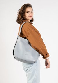 Graue Leder-Schultertasche mit einer geschwungenen Form, glatter Textur und einem einzigen verstellbaren Schulterriemen, getragen von einer Person in einem braunen Hemd.