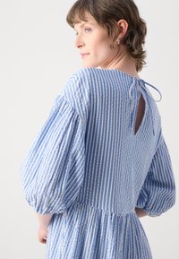 Lyseblå kjole med hvite horisontale striper, pufferm, og en bakre nøkkelhulldetalj knyttet med en sløyfe. Teksturert stoff, knelengdes design.