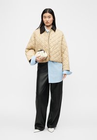 Γυναίκα με μπεζ μπουφάν με ραφές πάνω από μπλε φαρδιά πουκάμισο, μαύρο φαρδύ παντελόνι, λευκά μυτερά παπούτσια, κρατώντας μια μικρή κρεμ τσάντα χειρός.