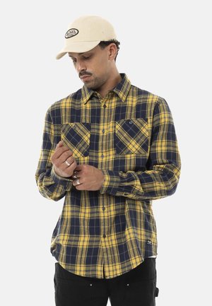 Homme ajustant la manche d'une chemise à carreaux jaune et marine, portant un pantalon noir et une casquette beige avec un logo à l'avant.