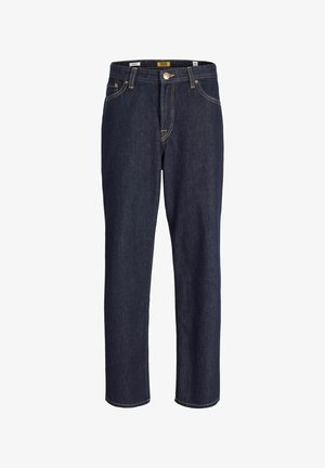 Persona con camisa beige abotonada sobre camiseta naranja quemado, pantalones rectos azul oscuro y zapatos negros con cordones, de pie frente a un fondo liso.