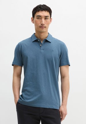 Mężczyzna stojący z rękami w kieszeniach, ubrany w niebieską koszulkę polo z krótkim rękawem i ciemne spodnie na białym tle.