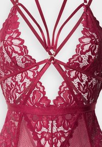 Μπορντό δαντελένιο εσώρουχο με περίτεχνα λουλουδένια σχέδια, ρυθμιζόμενους σατέν ιμάντες και κεντρική λεπτομέρεια με χρυσό κρίκο. Απαλή υφή από δίχτυ.