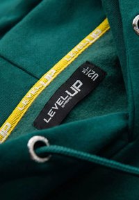Felpa verde con una texture morbida, caratterizzata da un'etichetta nera "LEVEL UP" e dettagli gialli con testo ripetuto lungo la cucitura interna. Dettaglio delle asole in metallo presente.
