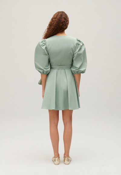 Rochie cu mâneci bufante dintr-un material moale de culoare verde mentă, cu talie ajustată, fustă evazată și o notă subtilă de strălucire. Asortată cu pantofi aurii. Fundal simplu.