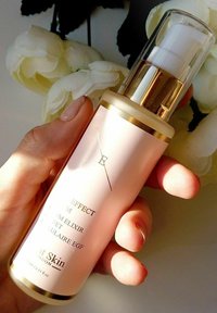 Różowa szklana butelka serum z złotymi akcentami. Pompka dozująca, etykieta "Effect M Serum Elixir", 70 ml. Trzymana w ręce, w tle delikatne kwiaty.