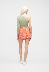 Γυναίκα με μακριά ξανθά μαλλιά στέκεται με την πλάτη, φορώντας πράσινο τοπ με ένα μανίκι, πορτοκαλί φο ύστα με λουλούδια και άσπρα αθλητικά παπούτσια σε απλό φόντο.