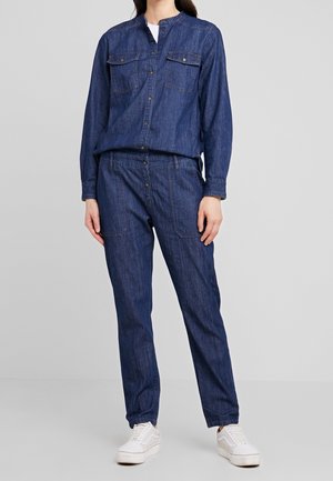 Femme portant une chemise en denim bleu foncé à boutons avec deux poches poitrine et un pantalon en denim assorti, associée à des baskets blanches.