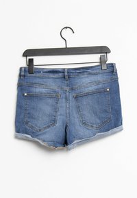 Farkkushortsit roikkumassa mustassa henkarissa. Väriltään keskiharmaansiniset, taitetulla helmalla ja kahdella takataskulla, joissa näkyy ompelusaumat.