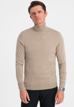 Un pull à col roulé beige en tricot léger, doté d'une silhouette ajustée et de poignets côtelés. Porté avec un pantalon bleu marine et une montre.