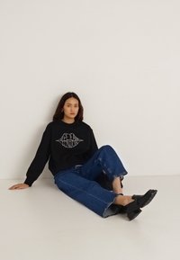 Μαύρο φούτερ με γραφικό τύπωμα, σε συνδυασμό με φαρδιά μπλε τζιν παντελόνια. Η εμφάνιση ολοκληρώνεται με μαύρα παπούτσια και τοποθετείται σε ουδέτερο φόντο.