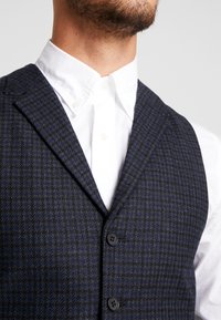 Chaleco de patrones de ajedrez en color azul marino con una mezcla de lana texturizada, con cuello de muesca y tres cierres de botón negro. Camisa blanca con cuello visible debajo.