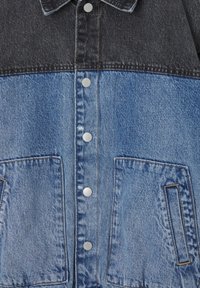 Veste en jean avec partie supérieure gris foncé et partie inférieure bleu clair, munie de boutons-pression argentés et de deux poches avant. Tissu texturé.