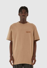 Beige oversized t-skjorte laget av bomull med korte ermer, med en liten brun logo på venstre side. Glatt tekstur, uformell design.