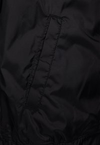 Czarna tkanina kurtki o gładkiej, lekko błyszczącej fakturze, z boczną kieszenią i widocznym przeszyciem. Brak wzorów i metalowych elementów.