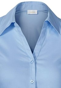 Camicia col celeste chiaro con colletto realizzata in tessuto liscio, con chiusura a bottoni sulla parte anteriore dotata di due bottoni e un'etichetta con la dicitura "atelier".