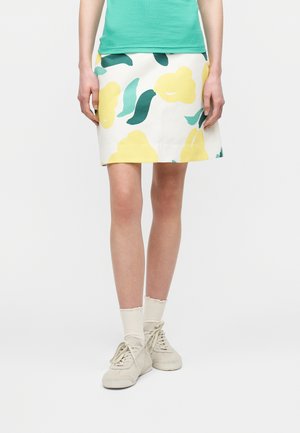 Γυναίκα που φορά λευκή φούστα με κίτρινο λουλουδάτο σχέδιο, πράσινο πάνω μέρος, λευκές κάλτσες και μπεζ αθλητικά παπούτσια, στέκεται μπροστά σε λευκό φόντο.