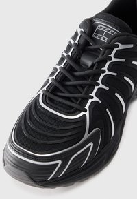 Scarpa sportiva nera con tomaia in rete testurizzata, design snello, accenti bianchi e sistema di allacciatura tradizionale. Suola spessa e imbottita.