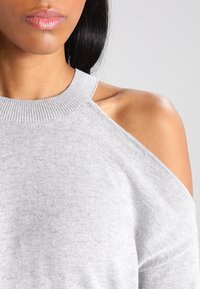 Haut en tricot gris clair avec une encolure dégagée et des découpes aux épaules. Texture douce avec des accents côtelés au niveau du col et des bords des manches.