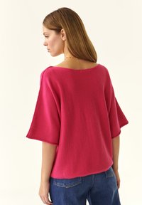 Maglione oversize rosa lavorato a maglia con scollatura ampia e maniche corte, caratterizzato da una texture morbida e da una vestibilità rilassata, abbinato a jeans blu.