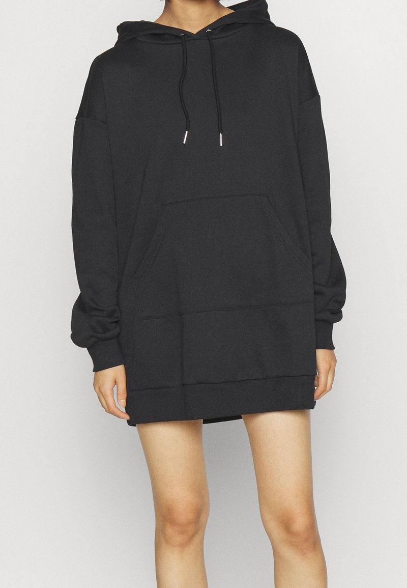 Personne portant une robe sweat à capuche noire surdimensionnée avec une poche avant et des cordons, debout devant un fond clair uni.