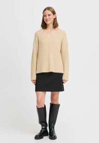 Beige gerippter Strickpullover mit V-Ausschnitt, oversized Ärmeln und rohem Saum, kombiniert mit einem schwarzen Minirock und knielangen schwarzen Stiefeln.