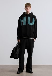 Μαύρο φούτερ με κουκούλα, με μεγάλο λογότυπο "HU" σε τιρκουάζ, συνδυασμένο με μαύρο παντελόνι, κρατώντας μια μαύρη τσάντα. Λείο ύφασμα, άνετη γραμμή.