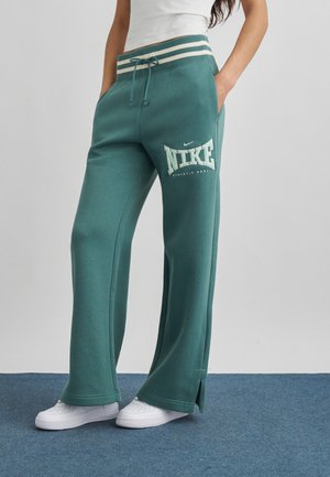 Teal Nike sweatpants med vide ben, med stribet linning og sideslids, båret med hvide sneakers og en hvid ærmeløs top.