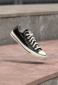 Czarny niskoprofilowy sneaker z białymi sznurowadłami i podeszwą, unoszący się nad różową betonową powierzchnią na tle szarej kamiennej ściany.