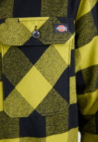 Grøn og sort ternet flannel skjorte med en stor brystlomme med knap og et mærketiket på lommens kant.