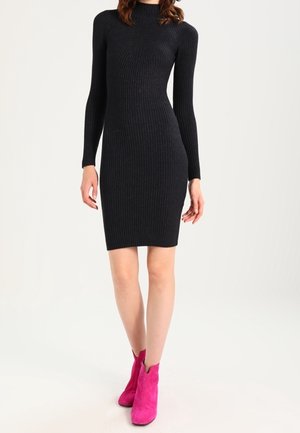 Frau trägt ein eng anliegendes schwarzes, geripptes Langarmkleid und leuchtend pinke Stiefeletten, steht vor einem schlichten hellen Hintergrund.