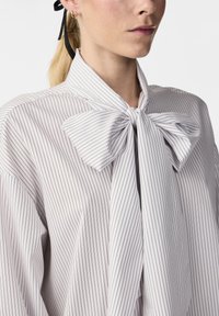 Blouse blanche à rayures avec un col à nœud oversized, présentant des lignes verticales noires, coupe ample et texture de tissu douce.