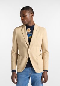 Beige blazer i bomull med notchad krage, två knappar och två framfickor. Bärs över en mörk mönstrad tröja med blå detaljer.