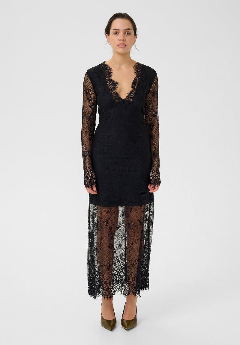 Robe en dentelle noire avec des manches longues, décolleté en V profond et motifs floraux complexes. Jupe transparente, longue jusqu'au sol, avec des détails en dentelle superposés.